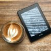 A Kindle resting on a wooden table beside a latte