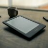 A Kindle e-reader resting on a wooden desk beside a cup of coffee